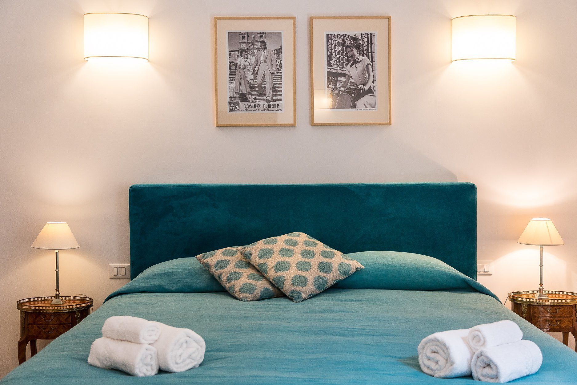Un lit bien fait, avec une couette et des draps de couleur turquoise, deux oreillers et des serviettes enroulées. Deux tableaux encadrés sont accrochés au mur, au-dessus du lit.