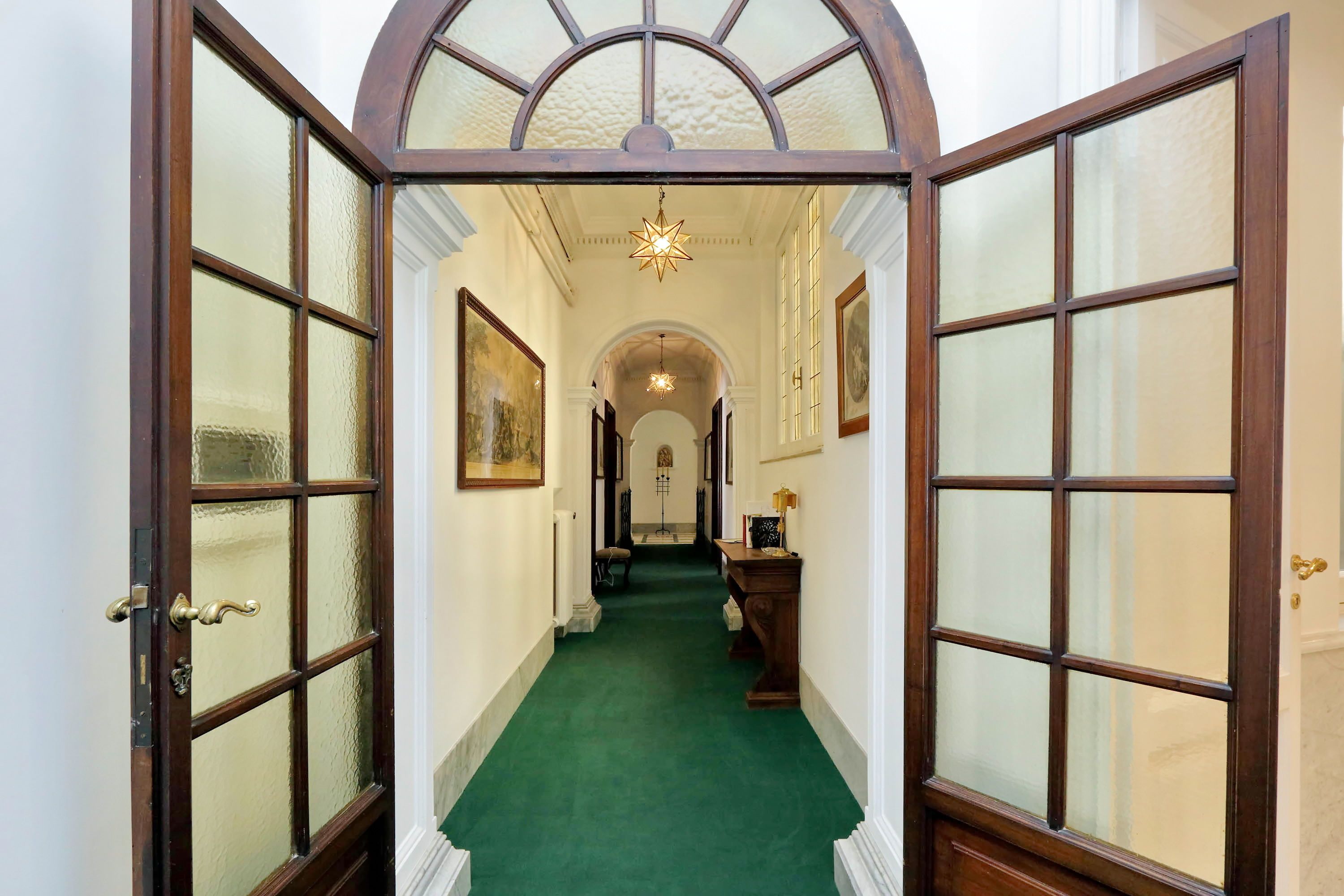 A hallway with a green carpet, wooden doors with glass panels, and framed artwork on the walls.