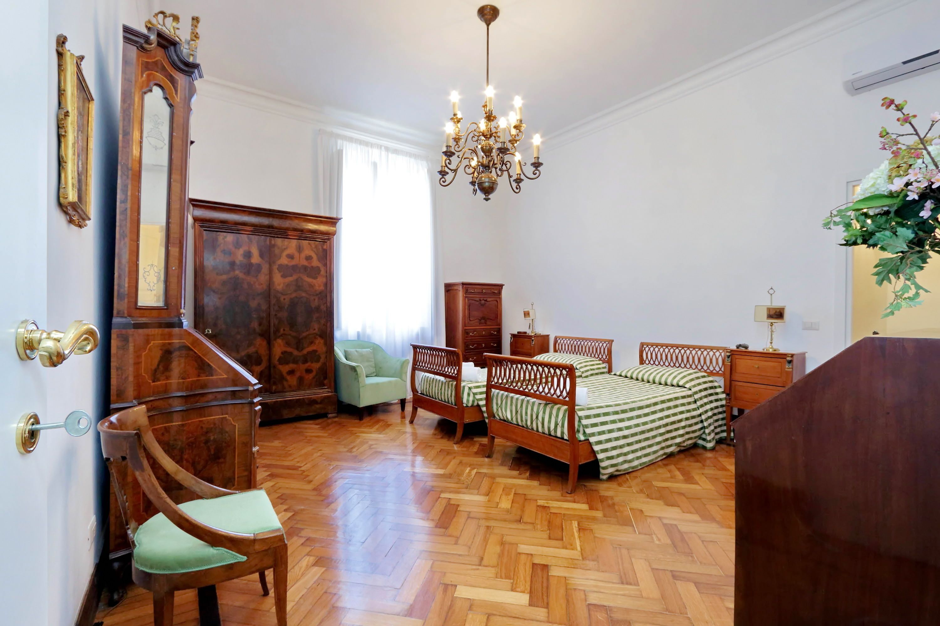 A bedroom with a wooden floor, a large mirror, a chandelier, a bed with green and white striped bedding, a wooden wardrobe, and a chair with a green cushion.