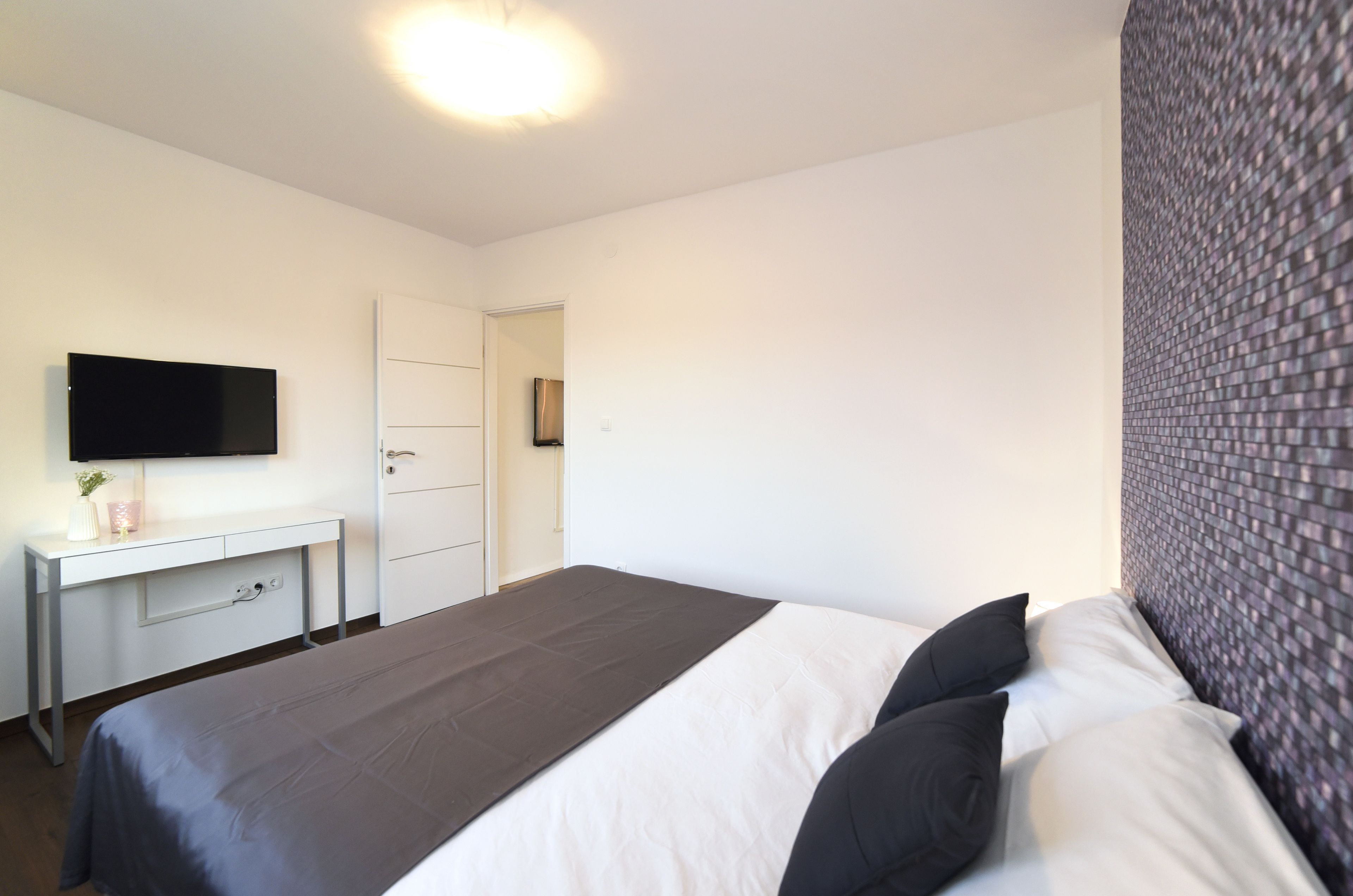 A bedroom with a bed, a desk, a TV, and a door.