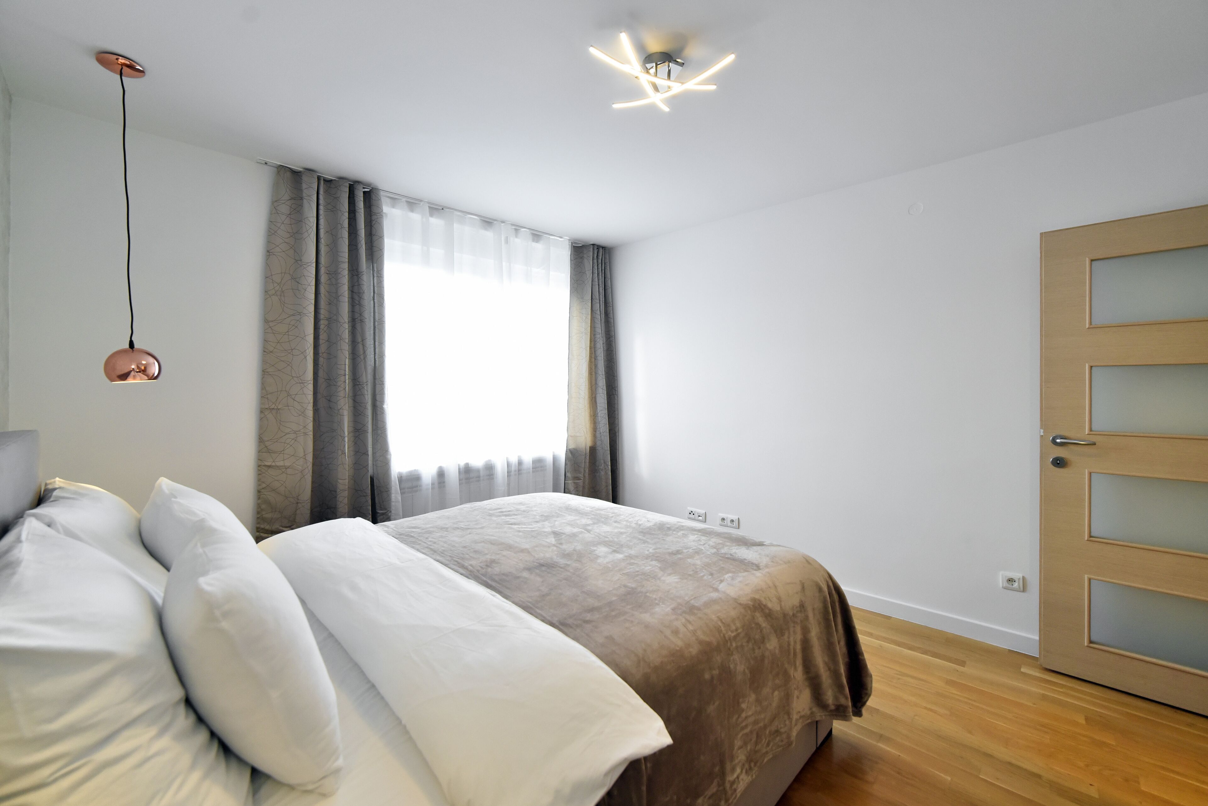 Una camera da letto con un letto, una lampada a sospensione, una finestra con tende e una porta in legno.