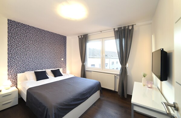 A modern hotel room with a large bed, a bedside table with a lamp, a flat-screen TV mounted on the wall, and a window with curtains.
