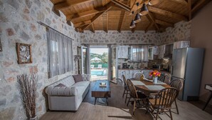A cozy living room with a sofa, dining table, and chairs, a kitchen with a refrigerator, and a view of a pool outside. - Roggaki Ionian Villas (Aktio-Vonitsa)