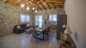 A spacious living area with a stone wall, wooden ceiling beams, a dining table with chairs, a sofa, and a kitchen area with a refrigerator and cabinets. - Roggaki Ionian Villas (Aktio-Vonitsa)
