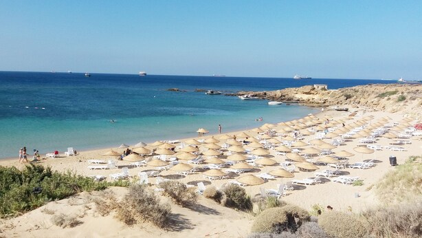 Une plage avec de nombreuses chaises longues et des parasols, un rivage rocheux et des bateaux sur l’eau.