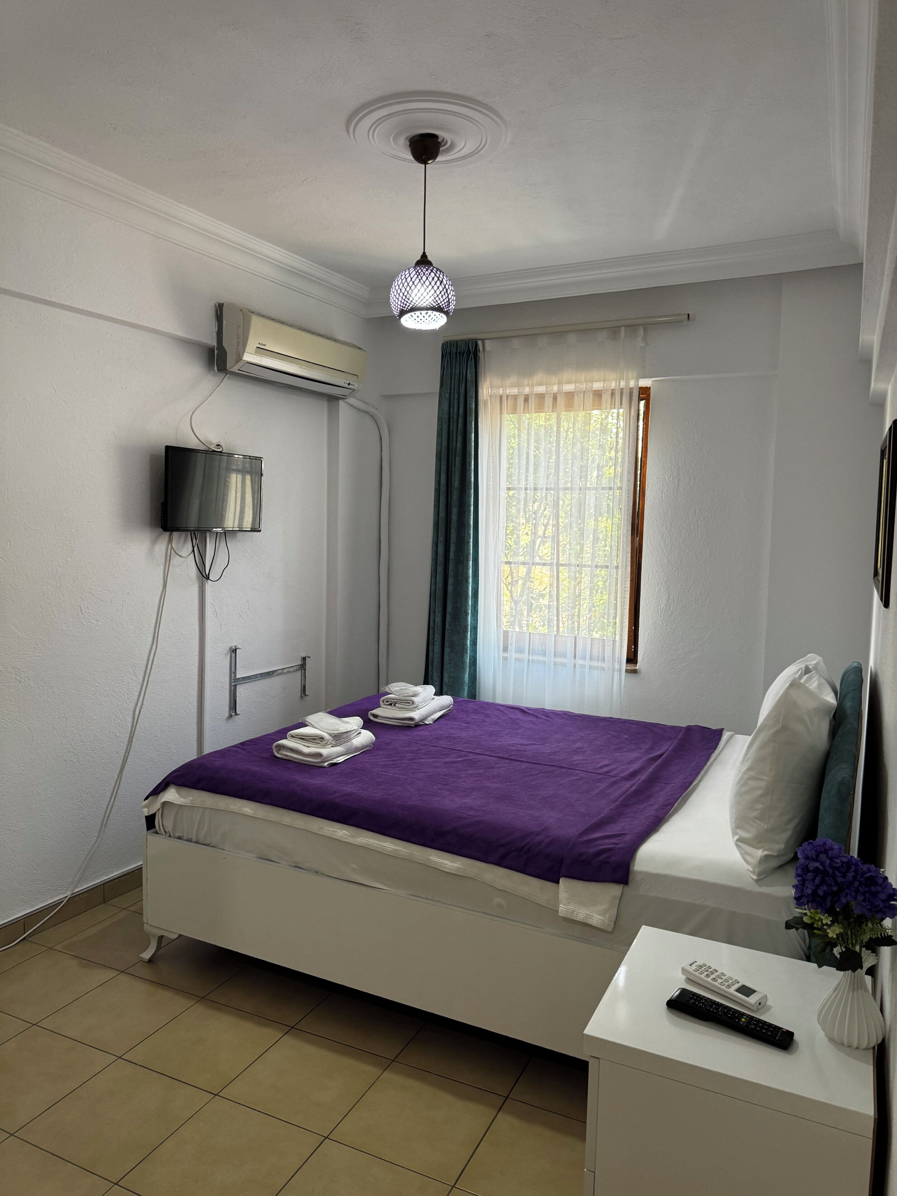 A hotel room with a bed, a bedside table, a TV, an air conditioner, and a window with curtains.