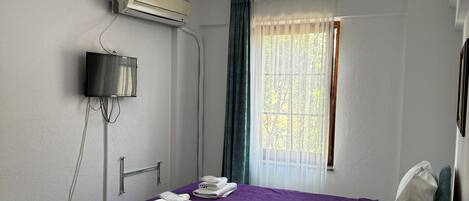 A hotel room with a bed, a bedside table, a TV, an air conditioner, and a window with curtains.
