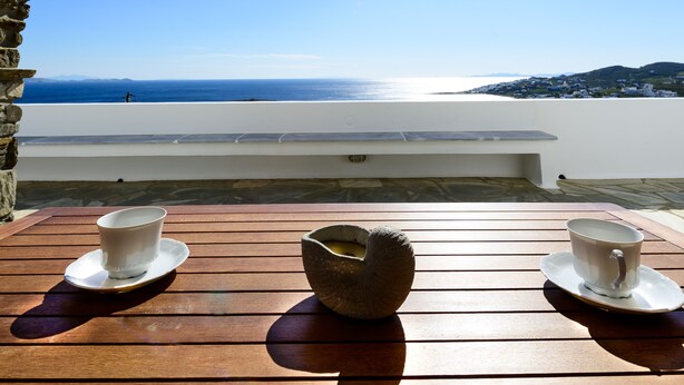 Une table en bois, sur laquelle reposent deux tasses et sous-tasses blanches, un bol en pierre, et d’où l’on a une vue dégagée sur la mer et les collines lointaines, le tout baigné par un soleil éclatant.