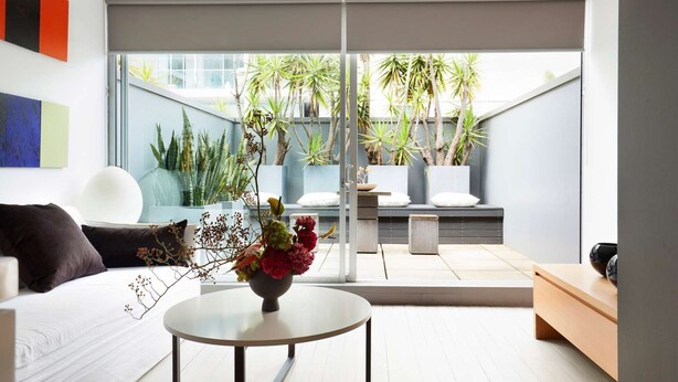 A modern living room with a sofa, a coffee table with flowers, and a view of a garden with various plants. - Apartment One by Contemporary Hotels is the quintessential glamorous Darlinghurst apartment for thos (Darlinghurst)