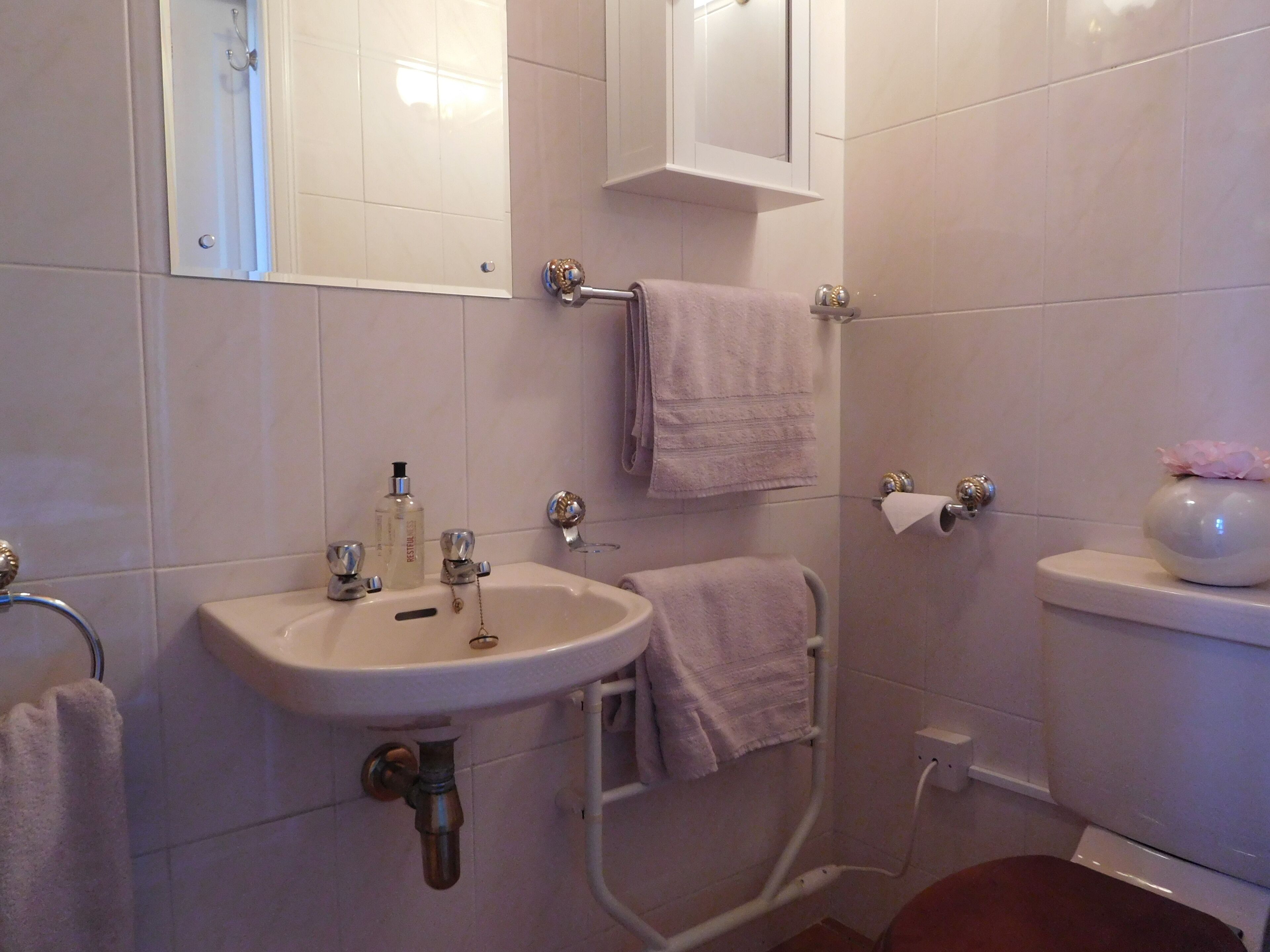 Un baño con lavamanos blanco, espejo y inodoro. Hay toallas colgadas en la pared y un florero sobre el inodoro.