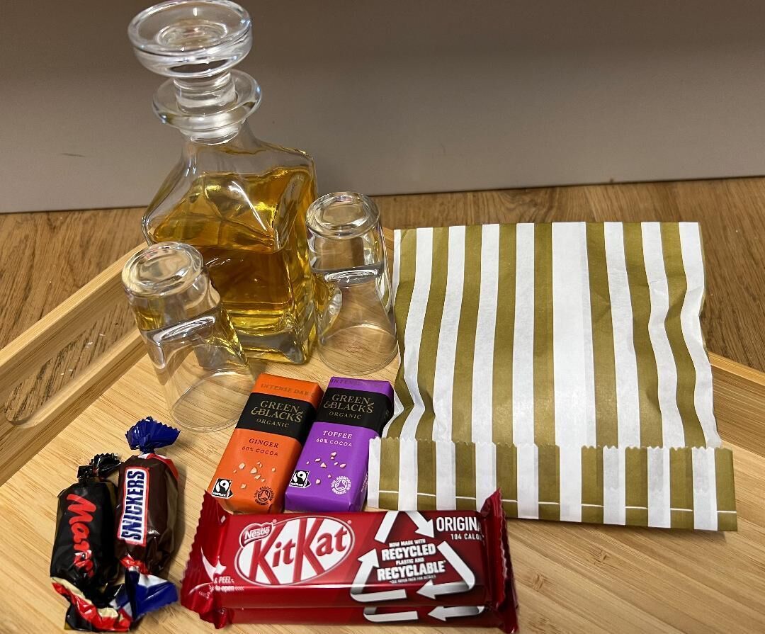 Un plateau en bois avec une bouteille en verre, deux verres à shooter, un torchon à rayures et diverses barres chocolatées, notamment des KitKat et des Snickers.