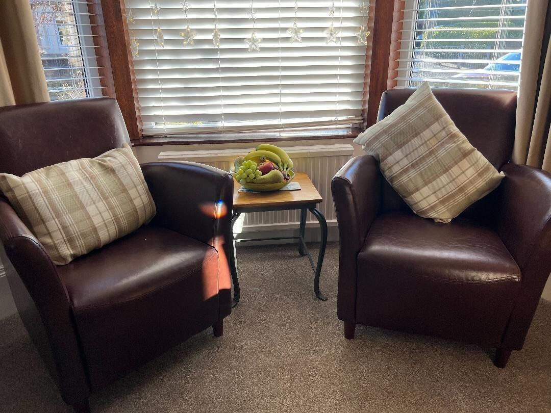 Dos sillones de cuero marrón con cojines de cuadros, una mesita de madera con un frutero, y una ventana con persianas.