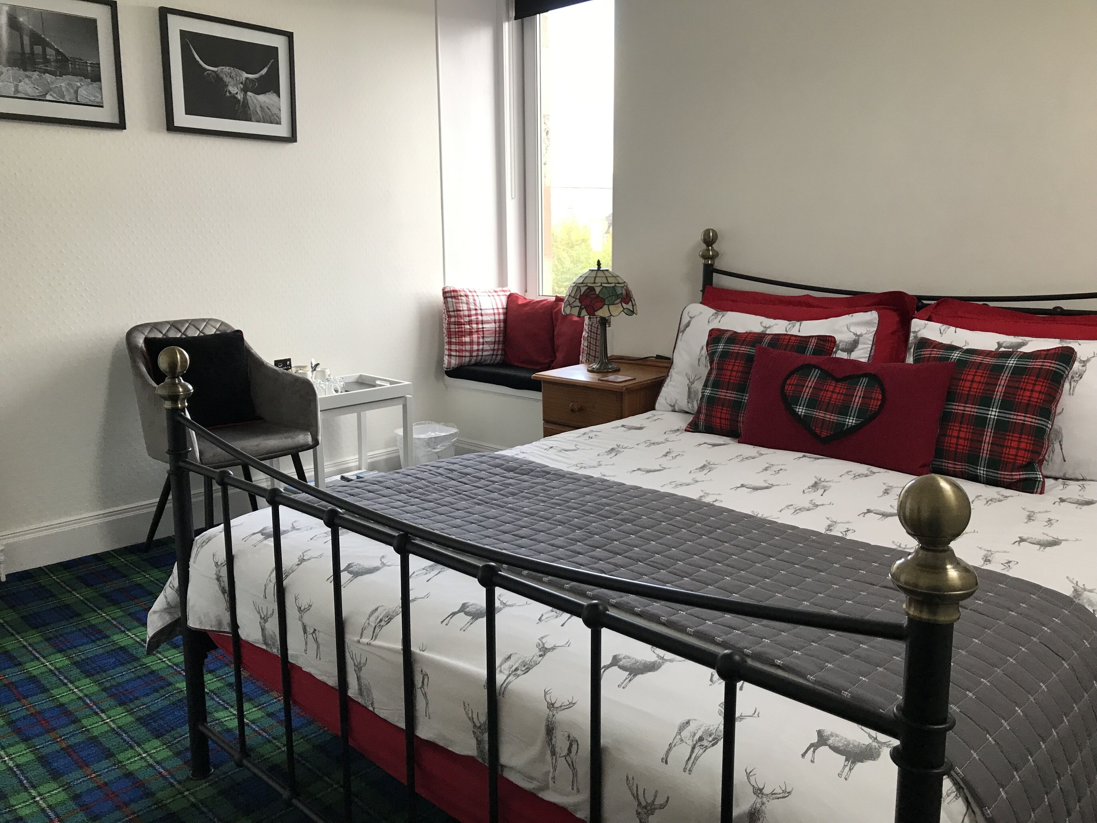 Un dormitorio con una cama que tiene una colcha con estampado de ciervo, un cabecero de metal negro, una silla con cojín negro, una mesita de noche con lámpara, y dos cuadros en la pared.