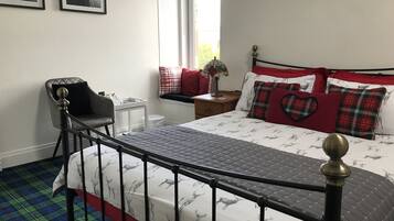 Un dormitorio con una cama que tiene una colcha con estampado de ciervo, un cabecero de metal negro, una silla con cojín negro, una mesita de noche con lámpara, y dos cuadros en la pared.