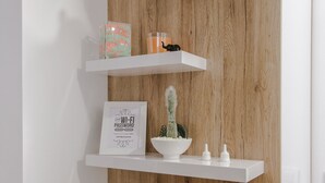Pared de madera con tres estantes blancos flotantes. El estante superior sostiene un florero blanco con plantas verdes y un pequeño objeto decorativo. El estante central tiene un cuadro con texto, un cactus pequeño y un cuenco blanco con adornos. El estante inferior tiene una planta en maceta.