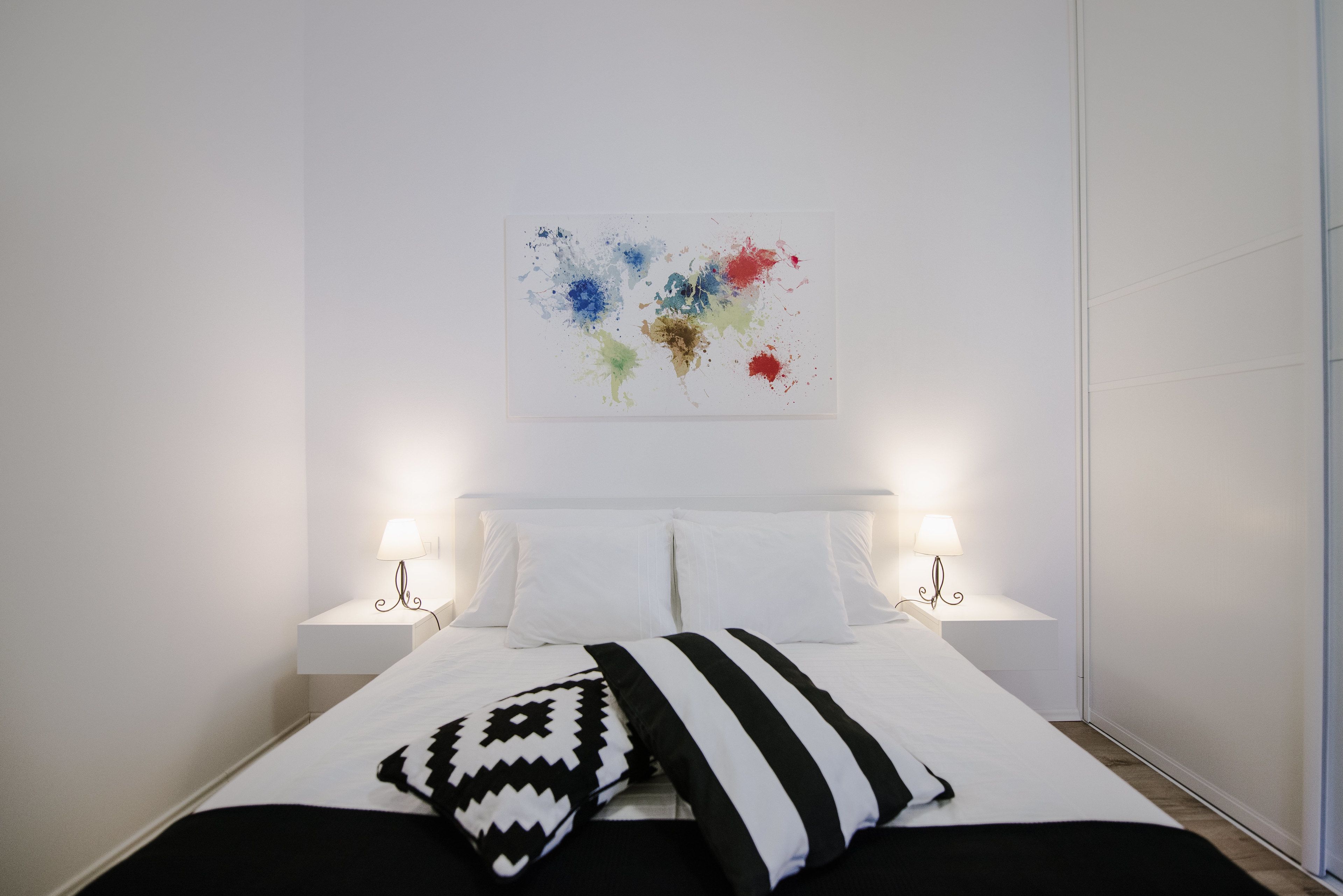 A neatly made bed with white linens, two bedside lamps, and a colorful abstract painting on the wall.