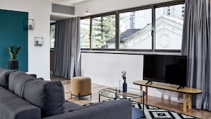 Una sala moderna con sofá, una mesa redonda, televisor y un ventanal con cortinas.