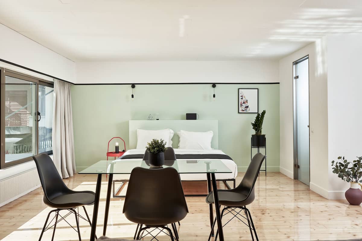 Quarto moderno com mesa e cadeiras de vidro, cama com roupa de cama branca e vista através de uma janela grande.