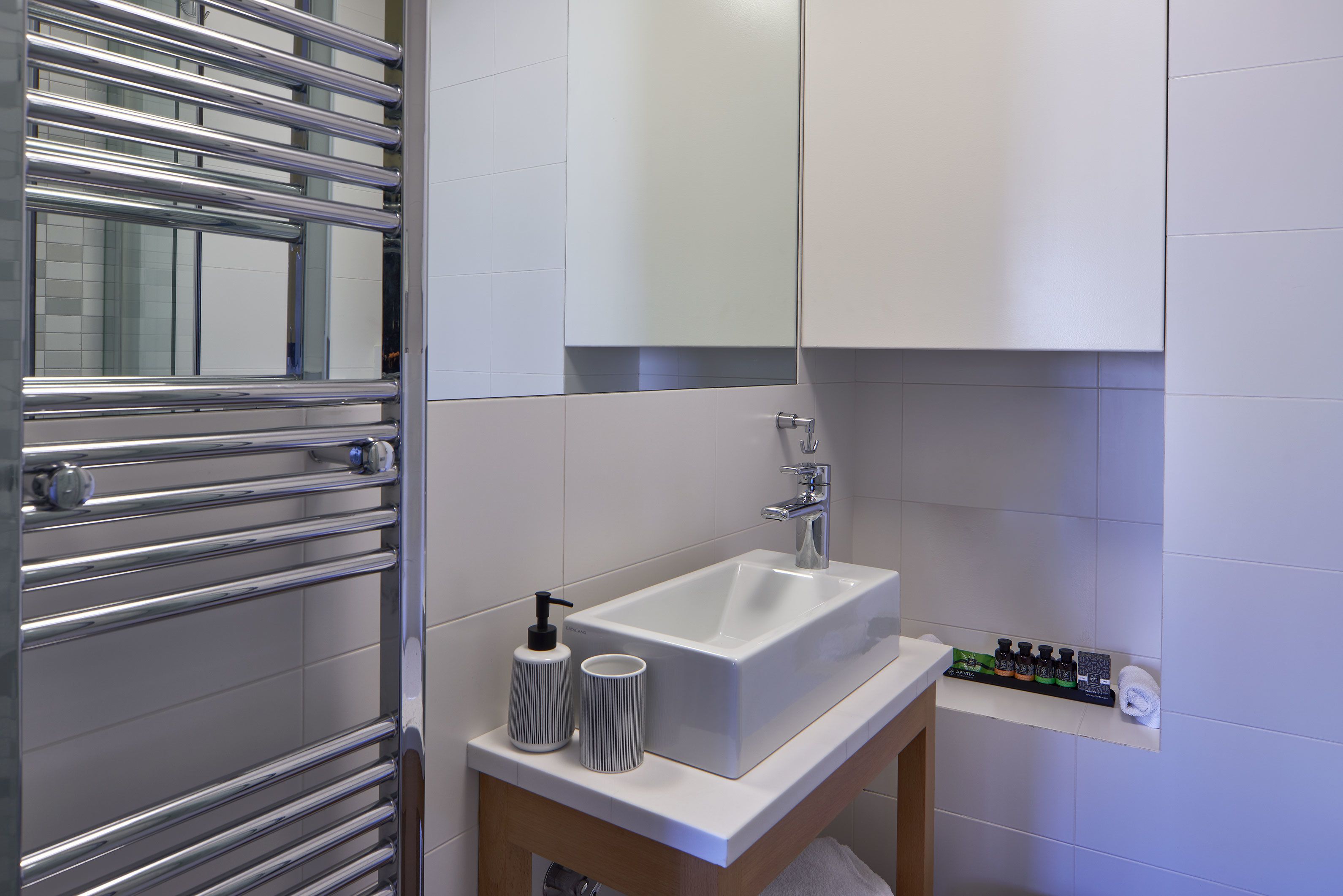 A modern bathroom with a white sink, chrome towel rack, and a shelf with toiletries.