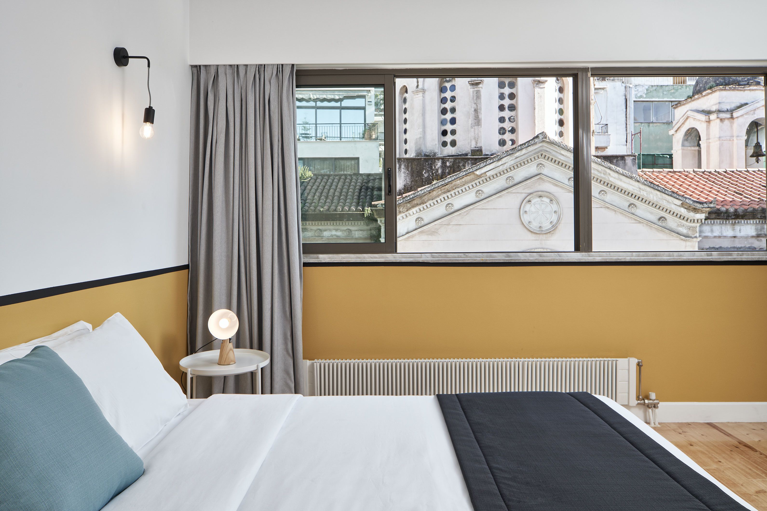 A hotel room with a bed, a nightstand with a lamp, a view of a building through a window, and a radiator.