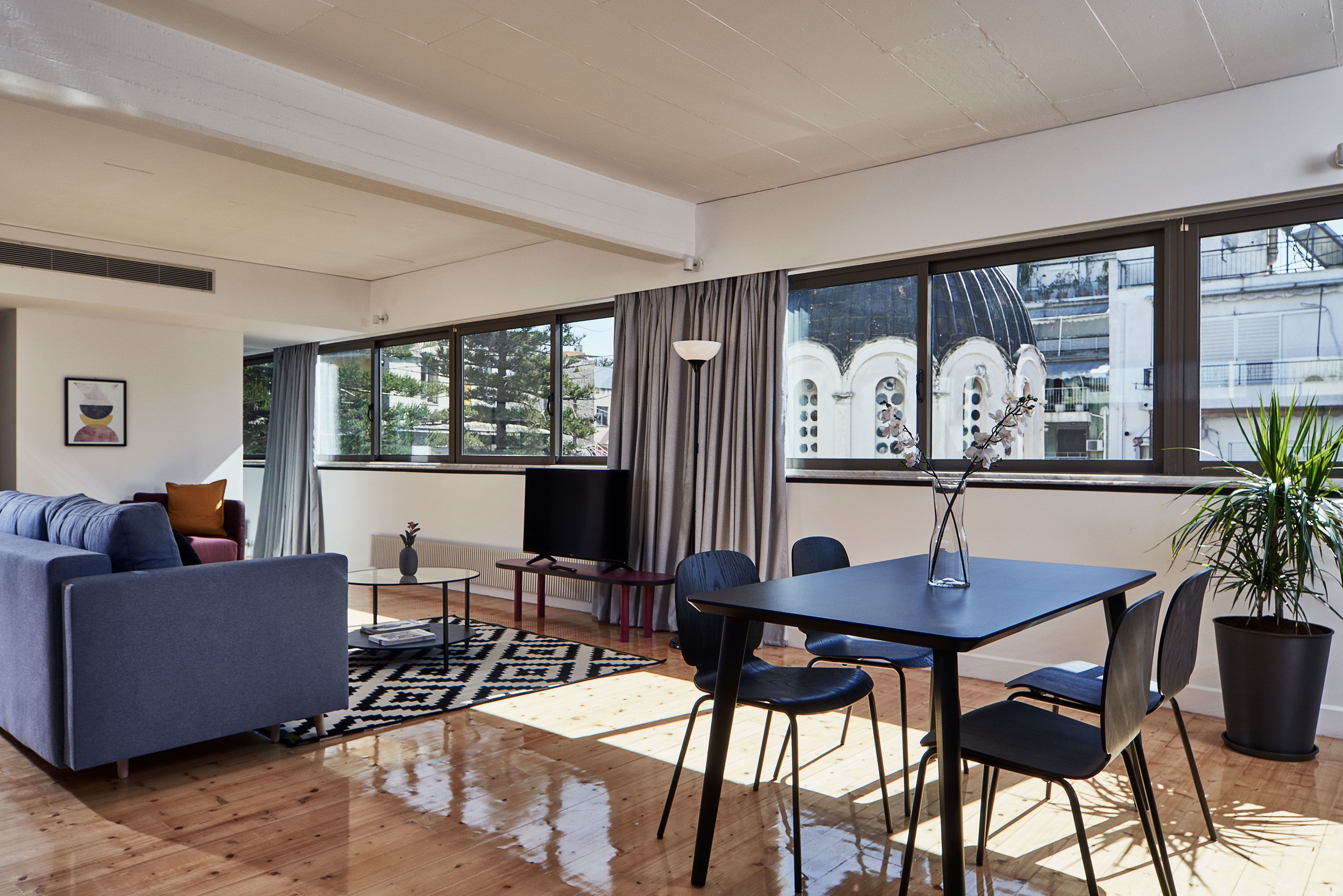 A modern living room with a sofa, a dining table, and chairs. There is a large window with curtains, a radiator, and a plant.