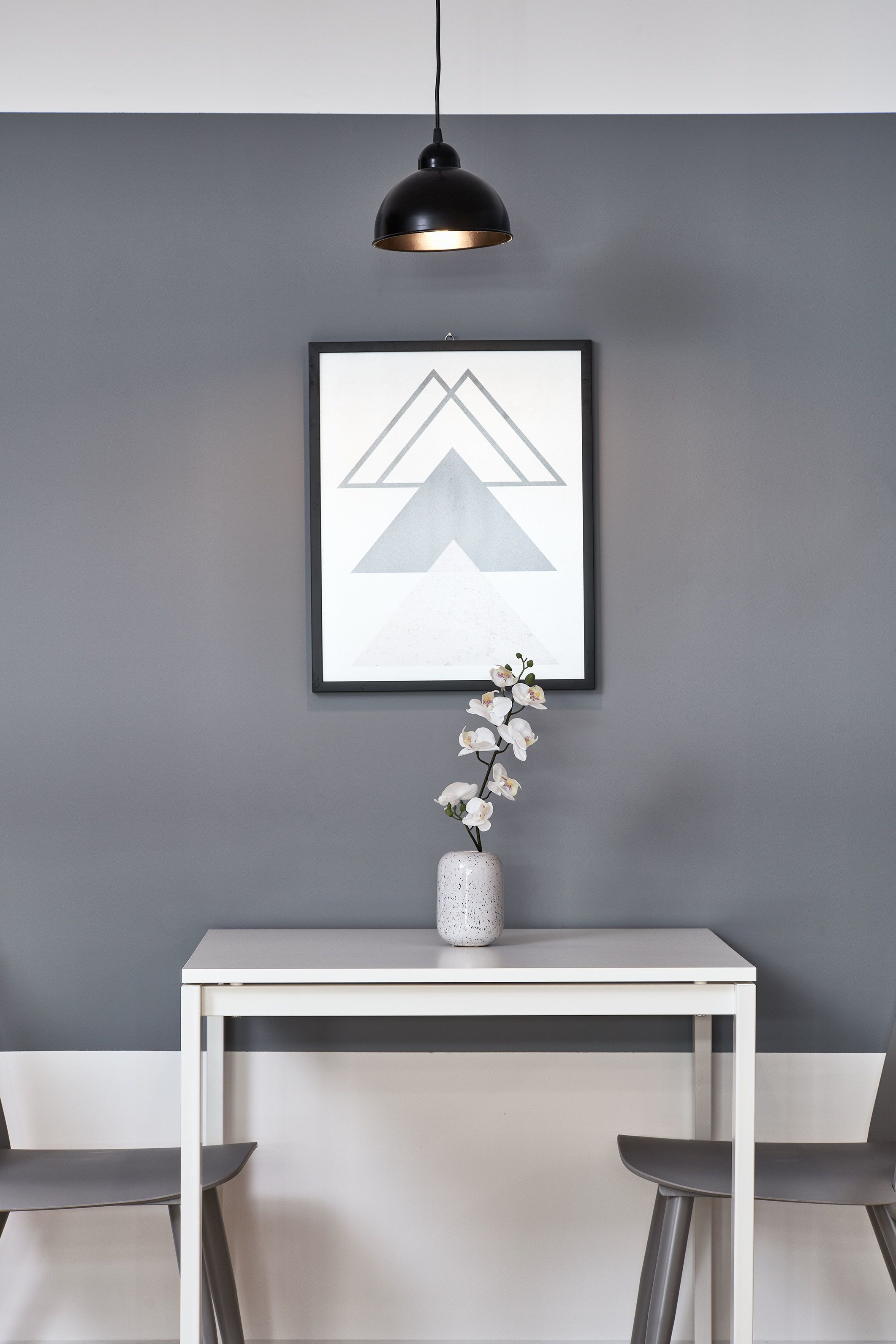 A modern room with a white table, two chairs, a framed abstract artwork, and a vase with white flowers.