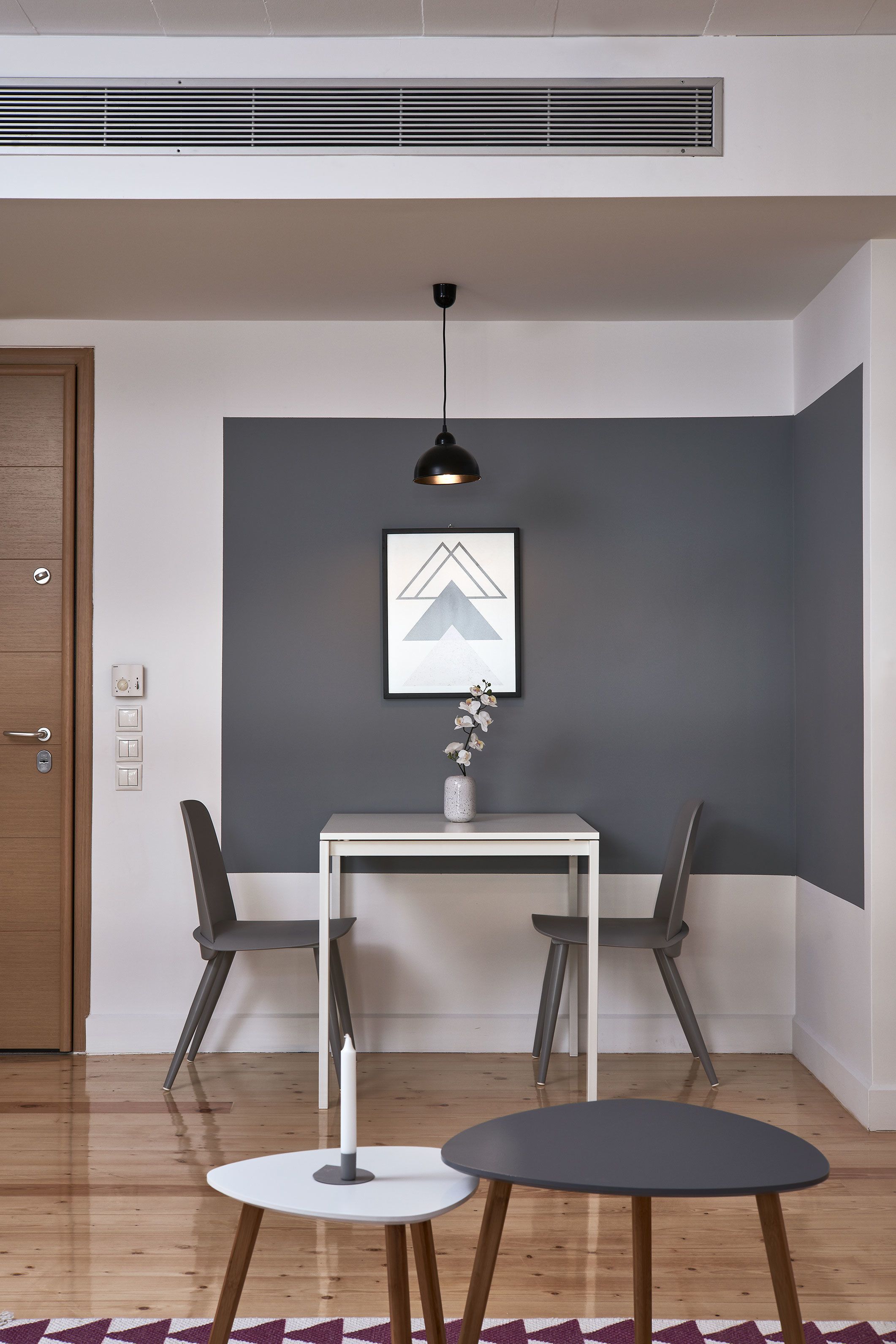 A modern living room with a white table, two grey chairs, a small round table, a pendant light, and a framed picture on the wall.