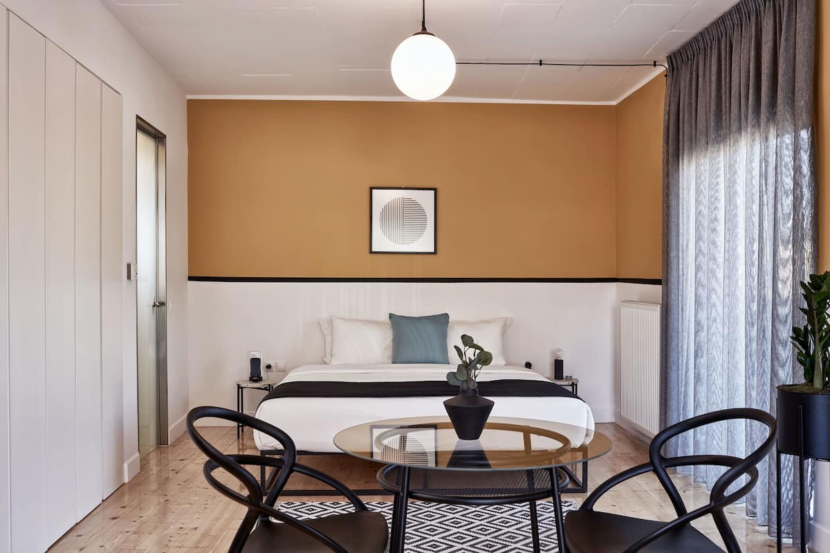 Quarto de hotel moderno com uma cama, uma mesa redonda de vidro, duas cadeiras, um vaso de plantas e uma obra de arte emoldurada na parede.