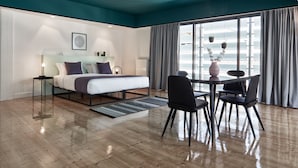 Una habitación de hotel moderna con una cama grande, una mesa de comedor con sillas, vistas de la ciudad a través de amplias ventanas y un techo pintado en color aguamarina.