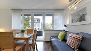 A modern living room with a grey sofa, wooden dining table, and chairs. A potted plant is on the table, and a window offers a view of buildings outside. - Vla Vla Apartments (Zagreb)