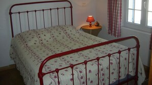 Una cama con cubrecama floral y estructura metálica roja, una mesita de noche con lámpara y una ventana con cortinas.