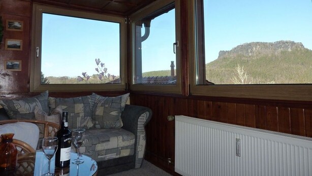 Una habitación acogedora con techo de madera, un banco acolchado y una mesa con una botella y vasos. Amplias ventanas ofrecen vistas a una colina.