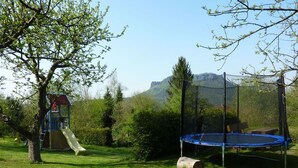 Un parque infantil con un tobogán y un trampolín en un área cubierta de césped, con árboles y una montaña al fondo.