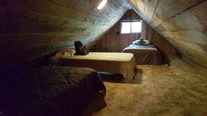 Une petite chambre mansardée, peu éclairée, avec deux lits, une fenêtre et un ours en peluche posé sur l’un des lits.