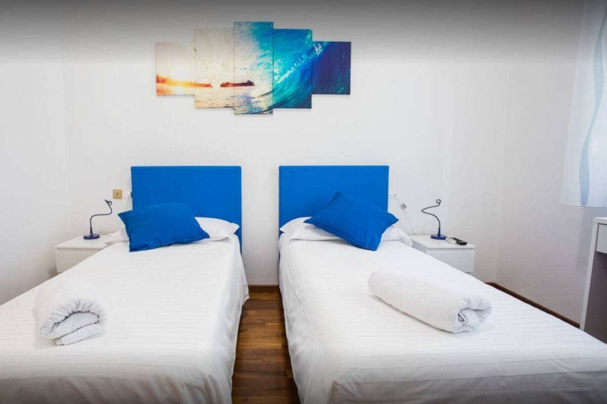 Habitación de hotel con dos camas, cada una con cabecera azul, ropa de cama blanca y una toalla blanca sobre la cama. Hay una obra de arte colgada en la pared sobre las camas.