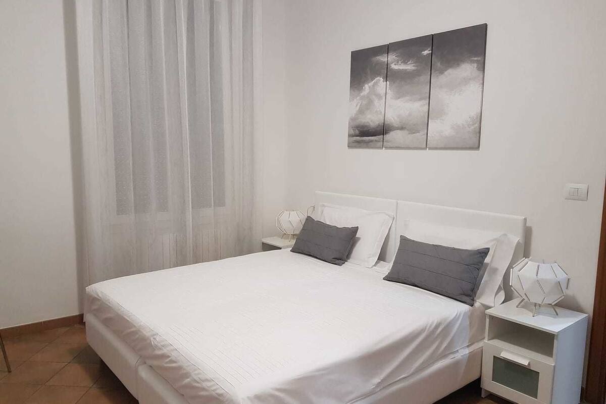 Un dormitorio con cama, dos mesitas de noche, un cuadro en la pared y una ventana con cortinas traslúcidas.