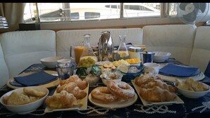 A breakfast table with pastries, fruits, and beverages, set in a room with white cushioned seating and a view of the outside. - Riti Yacht (Kalkara)