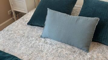 Una cama bien hecha con colcha blanca y azul turquesa, dos almohadas azul turquesa y una almohada gris. Un mesita de noche blanca con una lámpara y un dispositivo negro está a la izquierda.
