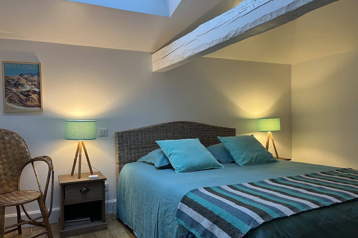 Une chambre à coucher avec un lit, deux lampes de chevet, une table de chevet, un fauteuil en osier et une affiche accrochée au mur.