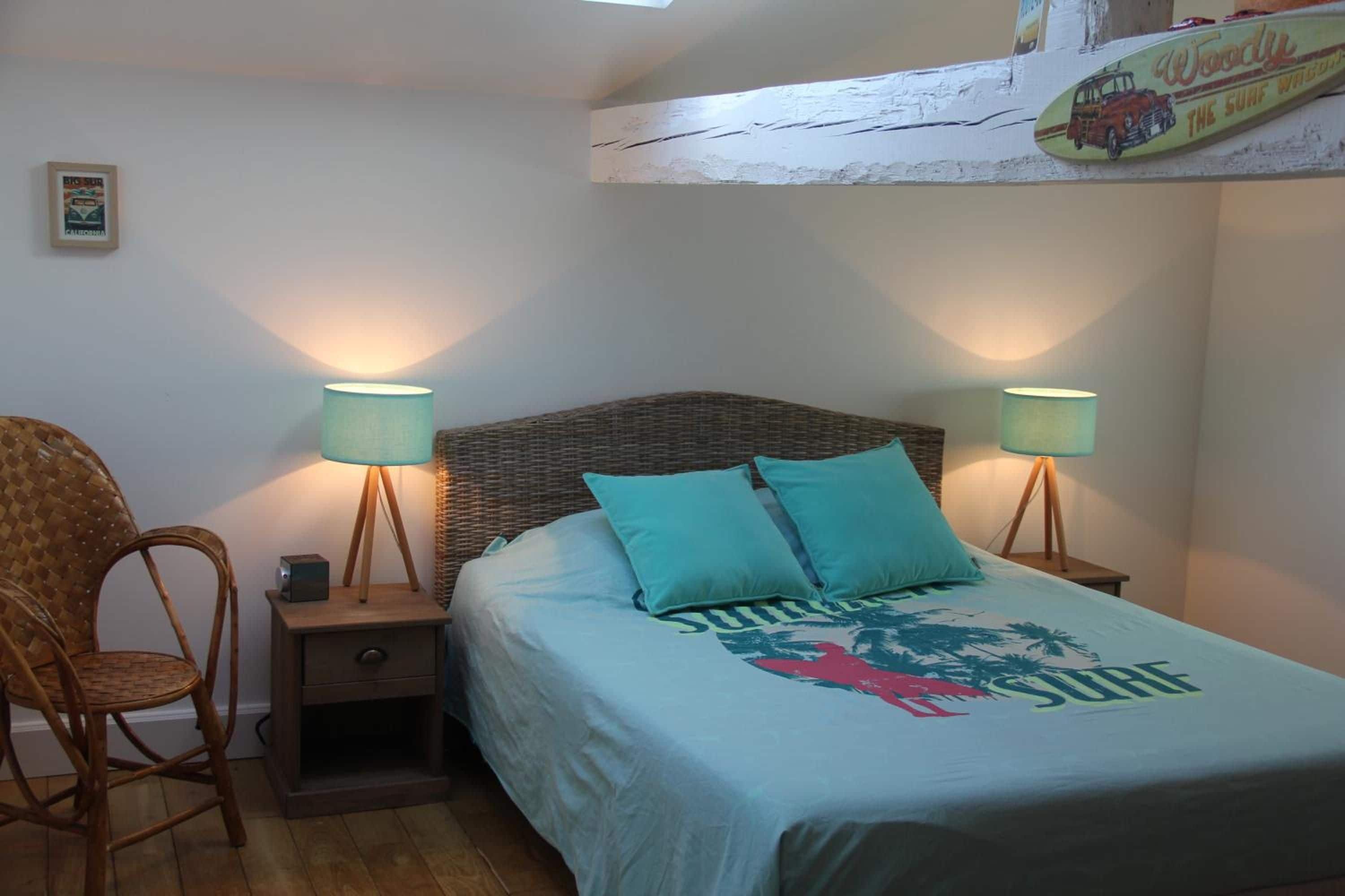 A bedroom with a bed, two bedside lamps, a wicker chair, and a small nightstand.