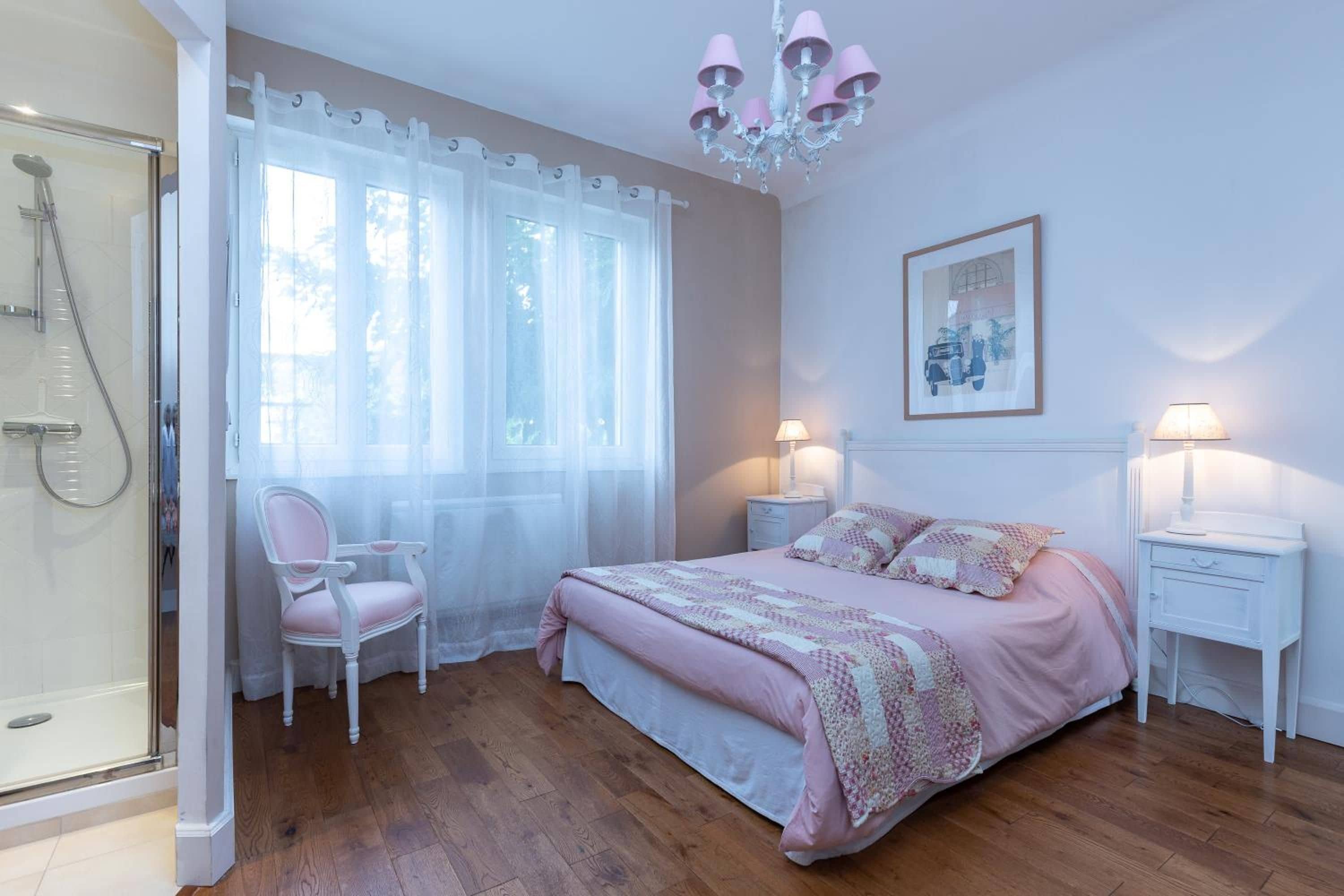 A bedroom with a bed, two bedside tables, a chair, a shower, and a chandelier.