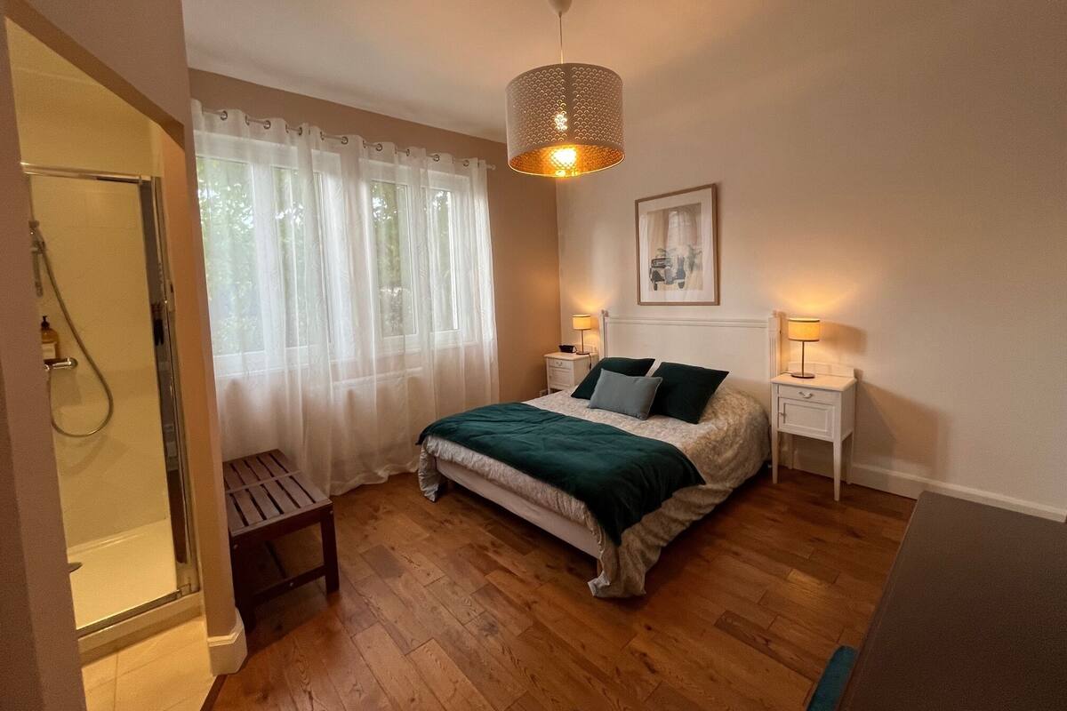 Une chambre à coucher comprenant un lit, des tables de chevet, un luminaire suspendu, une douche et une fenêtre avec des rideaux légers.
