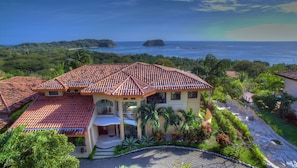 A large house with a tiled roof, surrounded by lush greenery and a view of the ocean.