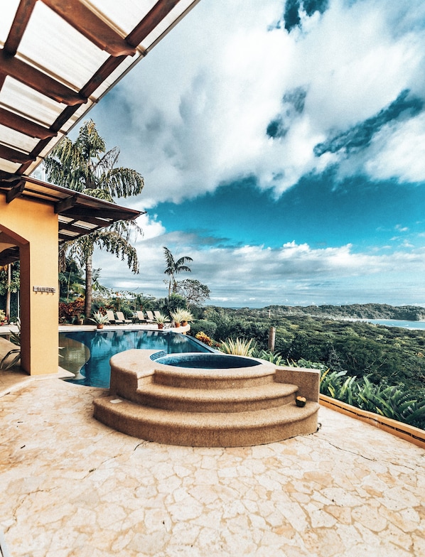 A pool area with a hot tub, lounge chairs, and a view of the ocean and hills.