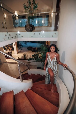 A woman sitting on a spiral staircase with a railing, surrounded by indoor plants and a dining area in the background.