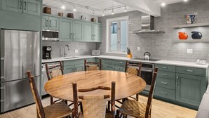 A modern kitchen with teal cabinets, a stainless steel refrigerator, a wooden dining table, and a brick backsplash.