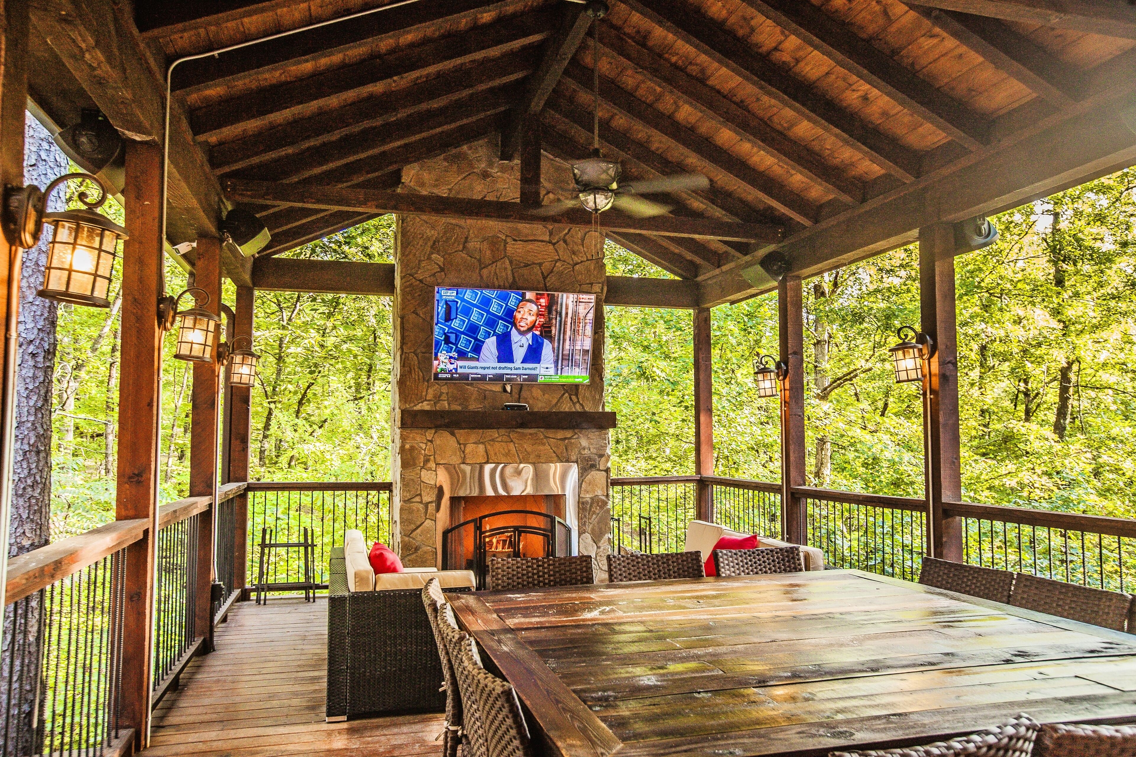 A covered outdoor seating area with a stone fireplace, a television mounted above it, and a view of the surrounding forest.