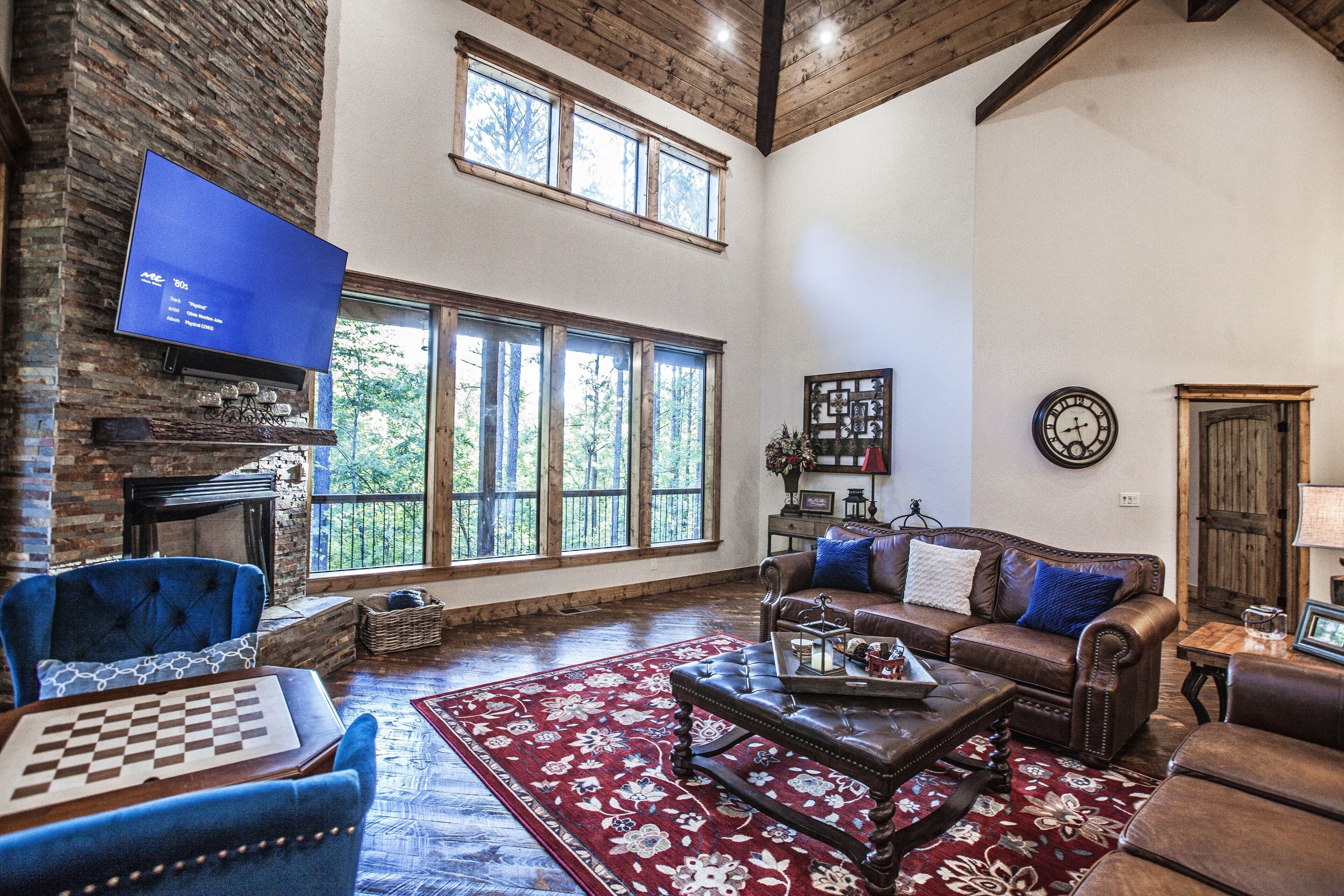 A spacious living room with a fireplace, a large TV mounted on a stone wall, a cozy seating area with a sofa and armchairs, and large windows offering a view of the outdoors.