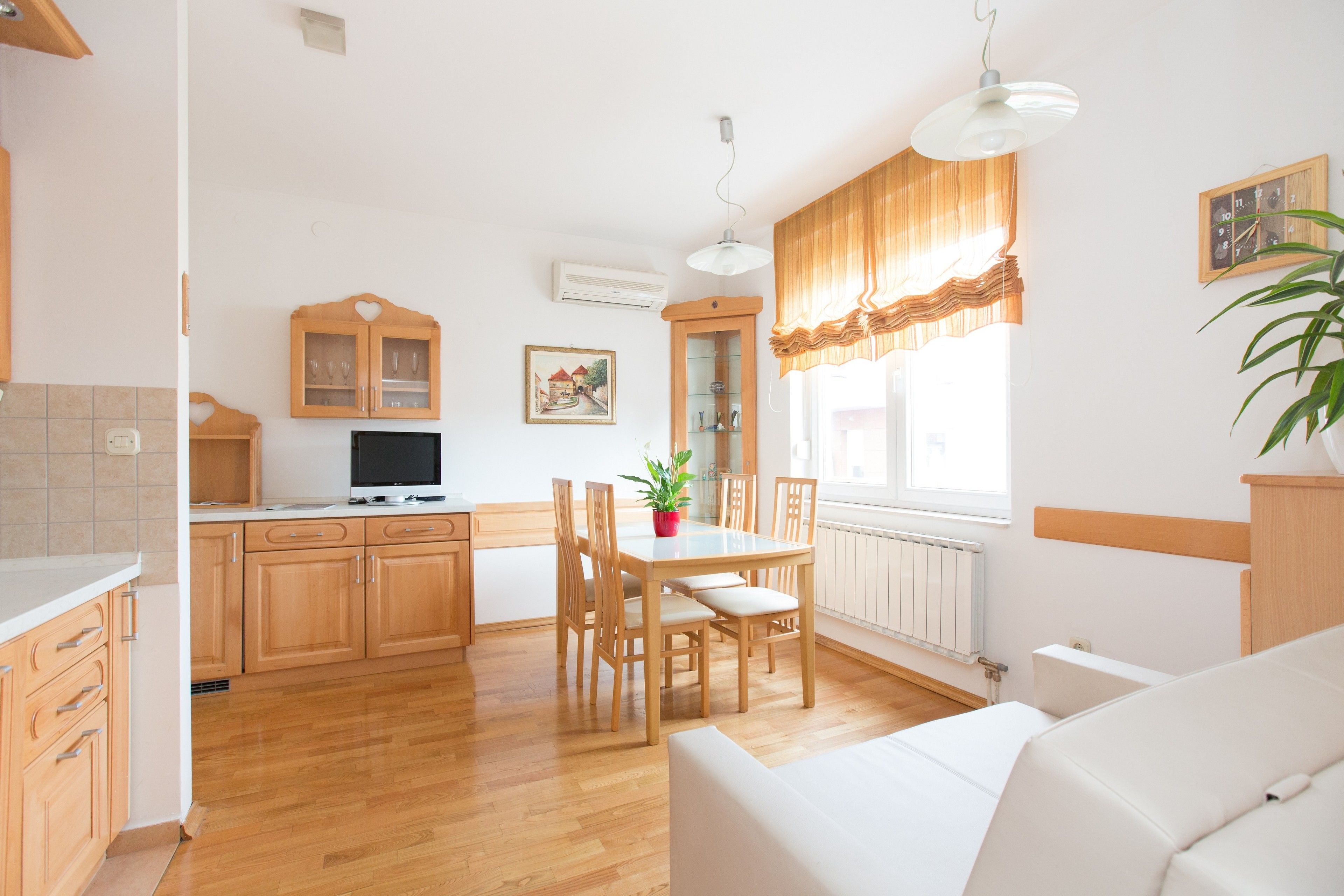 A modern kitchen with wooden cabinets, a dining table with chairs, a TV, and a potted plant.