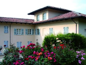 A building with a tiled roof, multiple windows with shutters, and a garden with various flowers. - Agriturismo La Casa In Collina (Canelli)